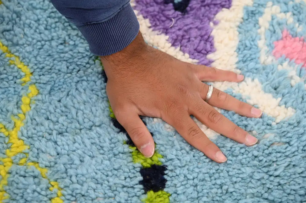 A hand with a silver ring rests on a plush moroccan handmade traditional azilal rug in light blue with multicolored patterns