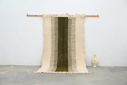 Beige and olive green moroccan handmade traditional azilal rug with woven texture and frayed edges hanging on wooden rod