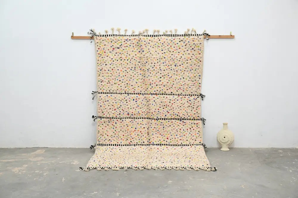 Beige moroccan handmade traditional azilal rug with colorful speckles and black bands hanging on wooden rod by decorbites