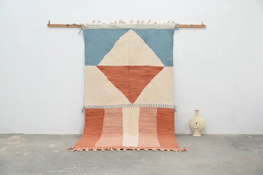 Moroccan handmade traditional azilal rug in soft blue terracotta and cream tones with fringed edges hanging on wooden rod