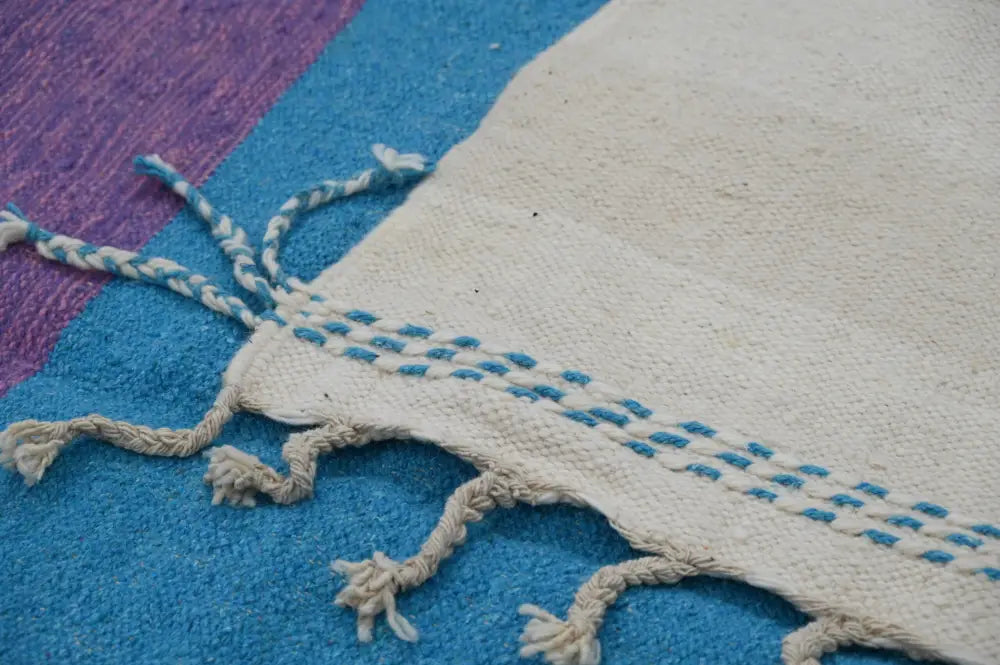 Cream-colored moroccan handmade traditional azilal rug with blue embroidered stitching and frayed braided tassels