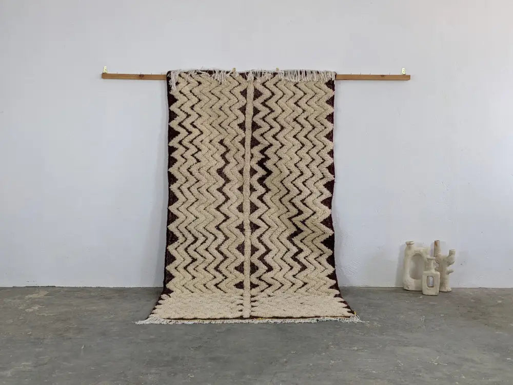 Beige and dark brown zigzag patterned moroccan handmade beni ouarain rug with fringed edges hanging on wooden rod