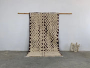 Beige and dark brown zigzag patterned moroccan handmade beni ouarain rug with fringed edges hanging on wooden rod