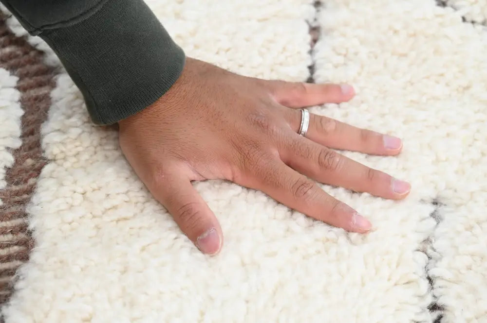 A hand resting on a plush cream beni ouarain rug with textured brown and black geometric stripes by decorbites