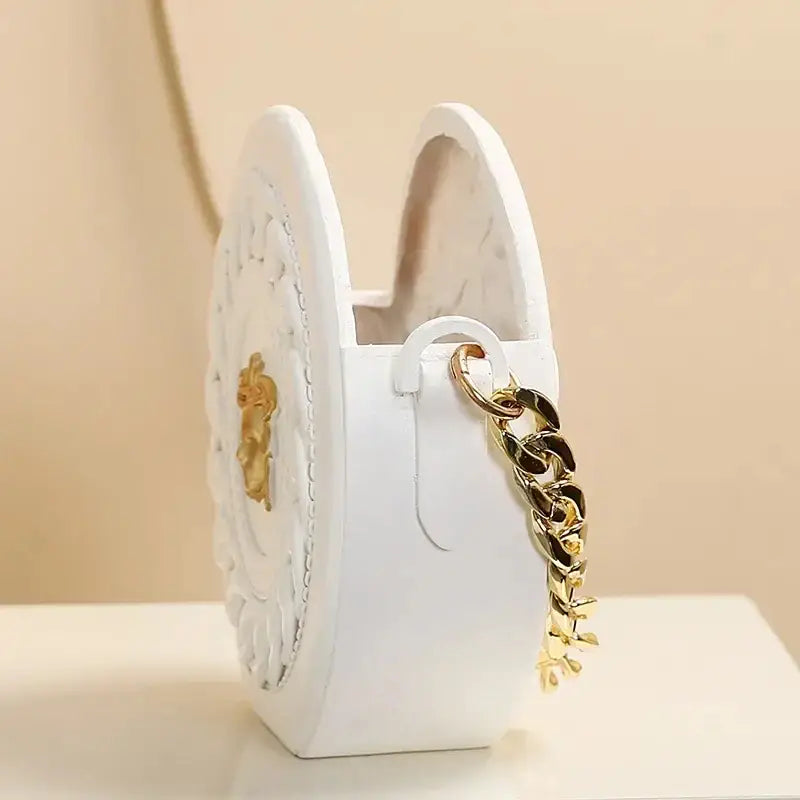 White circular handbag with gold-tone chain strap and embossed floral design next to nordic style ceramic flower vase decoration