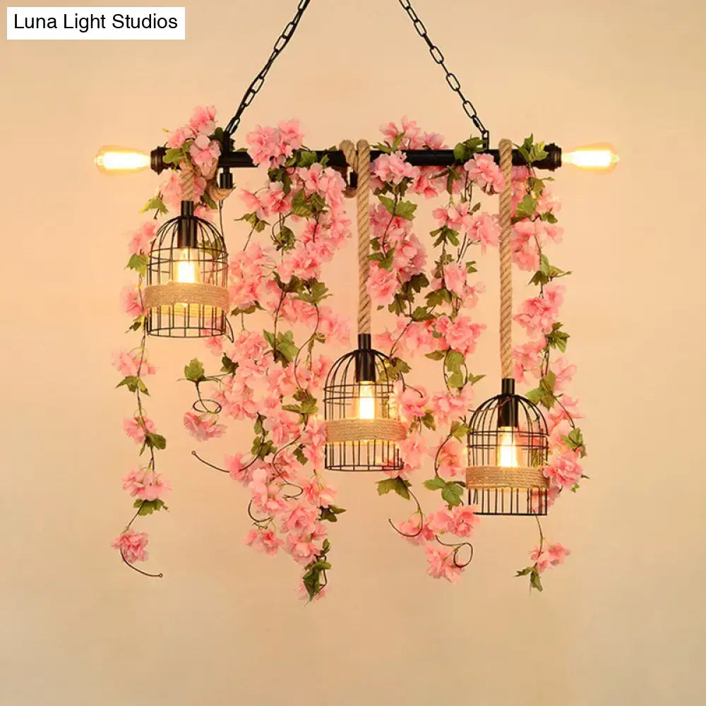 A rustic black metal chandelier with three birdcage lights featuring cascading pink floral vines for restaurants