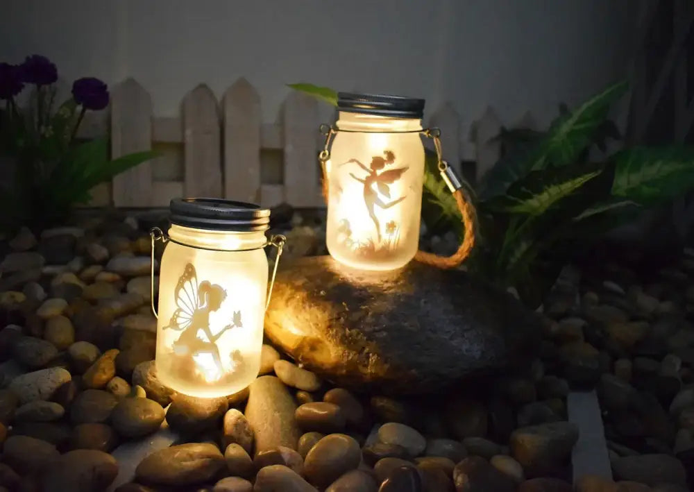 Two illuminated glass solar mason jar lanterns with fairy silhouettes, black lids, and metal handles by DecorBites™