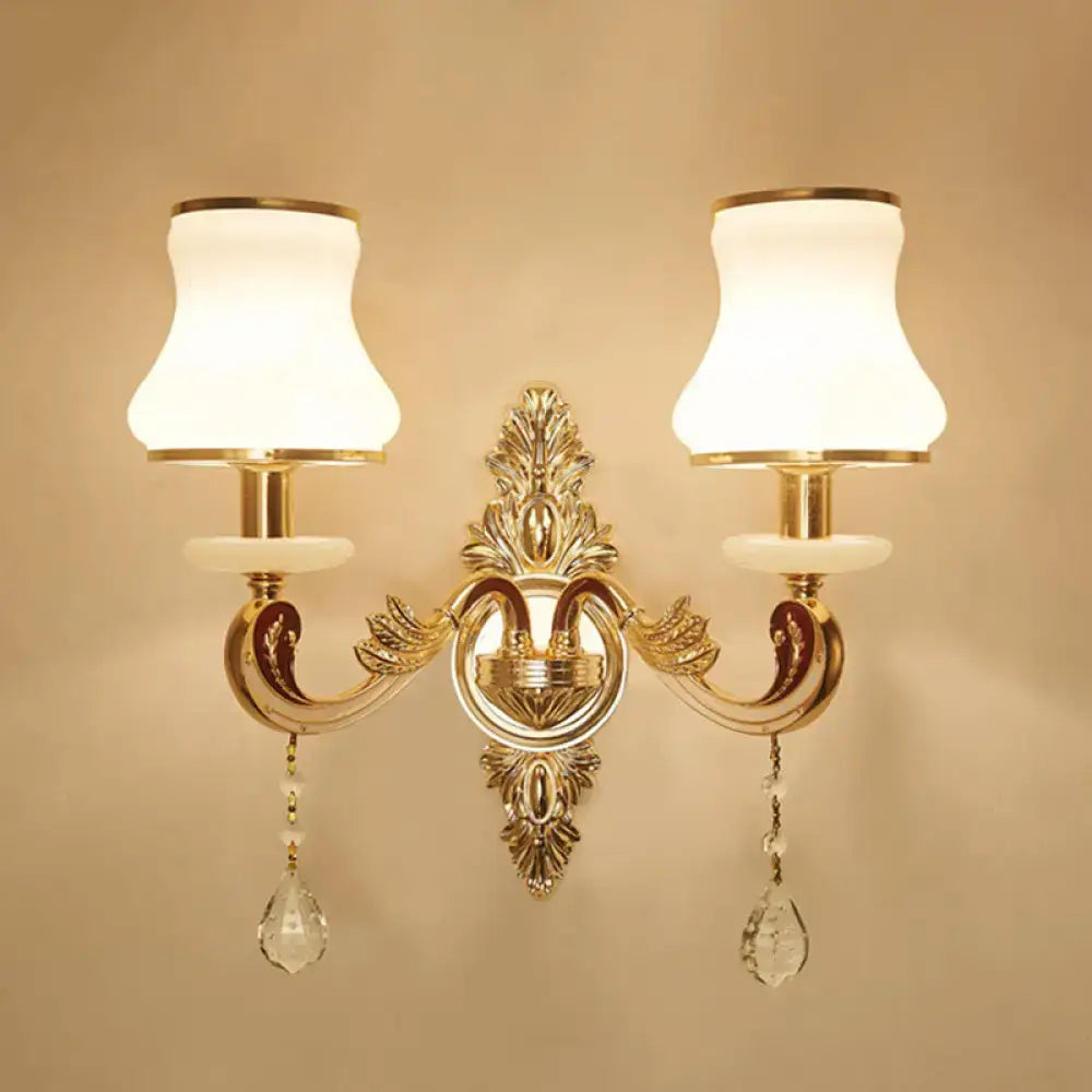 Elegant gold scrolled arm wall sconce with two frosted white lampshades and crystal teardrop pendants for bedroom use