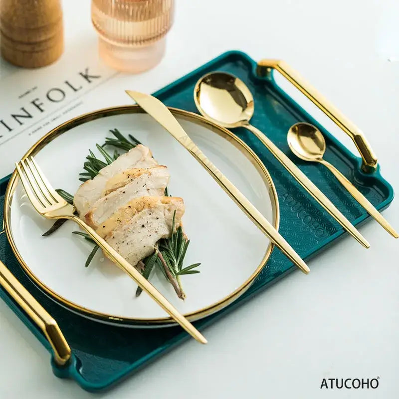 Elegant gold stainless steel cutlery set with sleek modern handles on teal tray beside white plate for dining room use