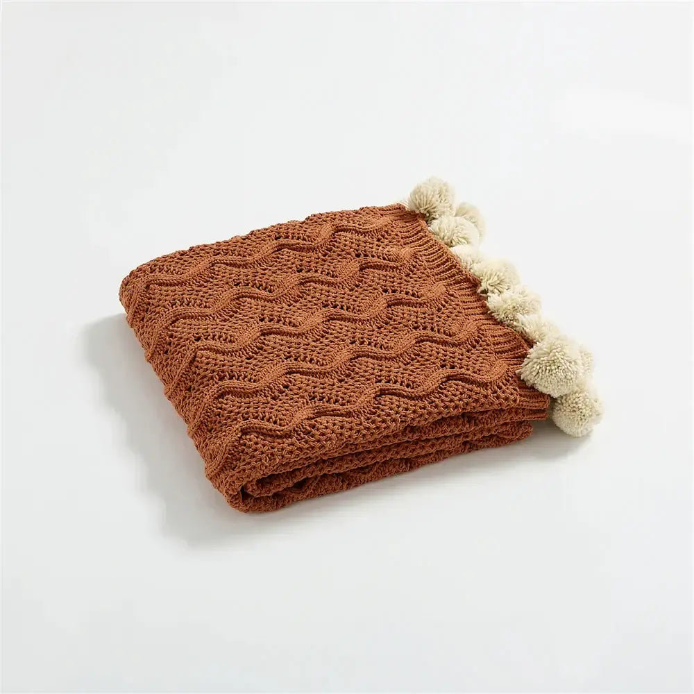Folded rust-colored knitted throw blanket with wavy cable pattern and cream pom-poms for decorbites elegant chenille decor
