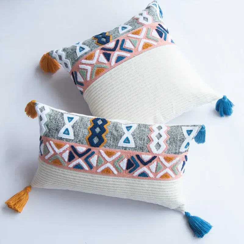 Rectangular embroidered blue tufted pillow cover with geometric patterns in gray, orange, and pink on white base with tassels