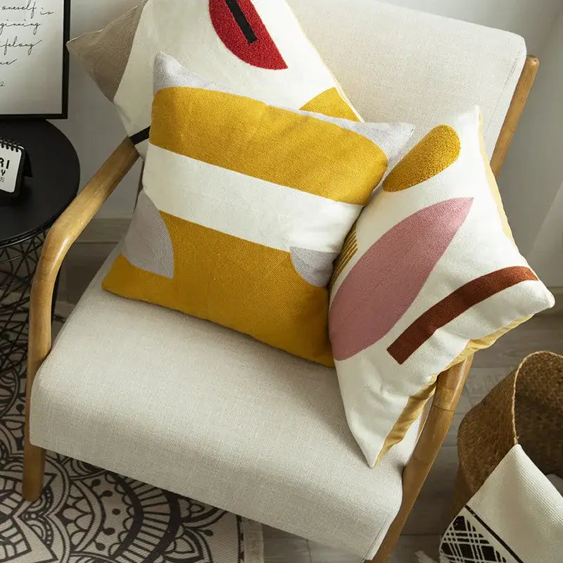 Two embroidered cushion covers with bold geometric patterns in mustard yellow, white, pink, and brown tones on armchair