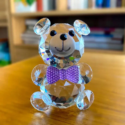 A faceted clear crystal teddy bear figurine with black button eyes and a purple jeweled bow tie for bedroom decor