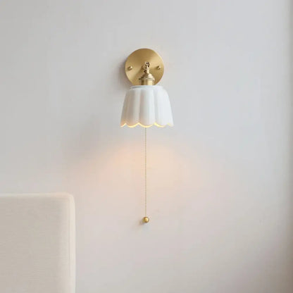 Delicate white ceramic wall light fixture with scalloped edge and gold pull chain for elegant bedroom lighting