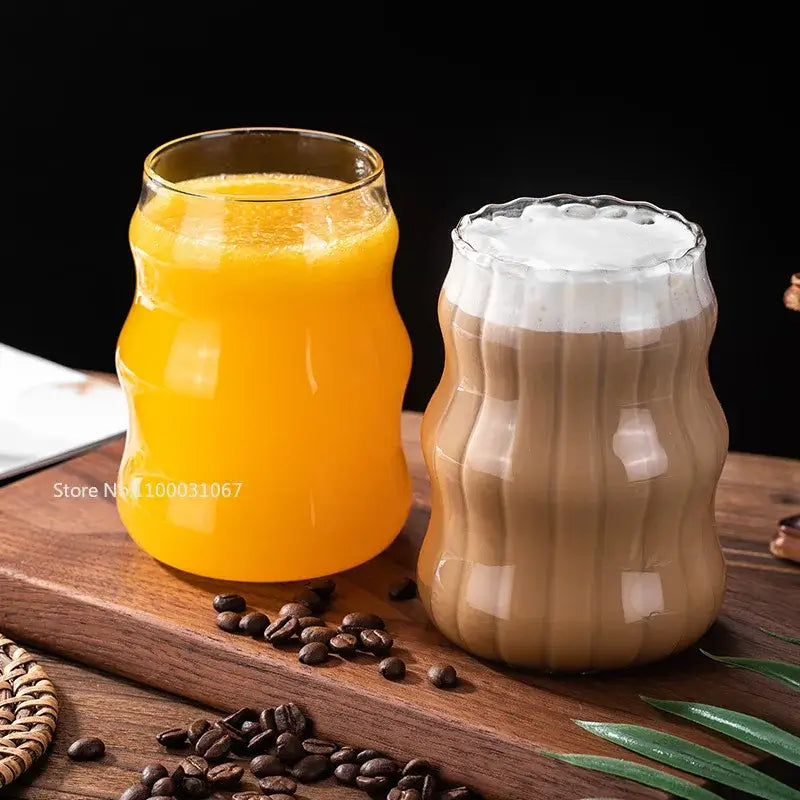 Two clear ribbed glass coffee mugs with ergonomic wavy design one with orange juice and one with latte on wood surface