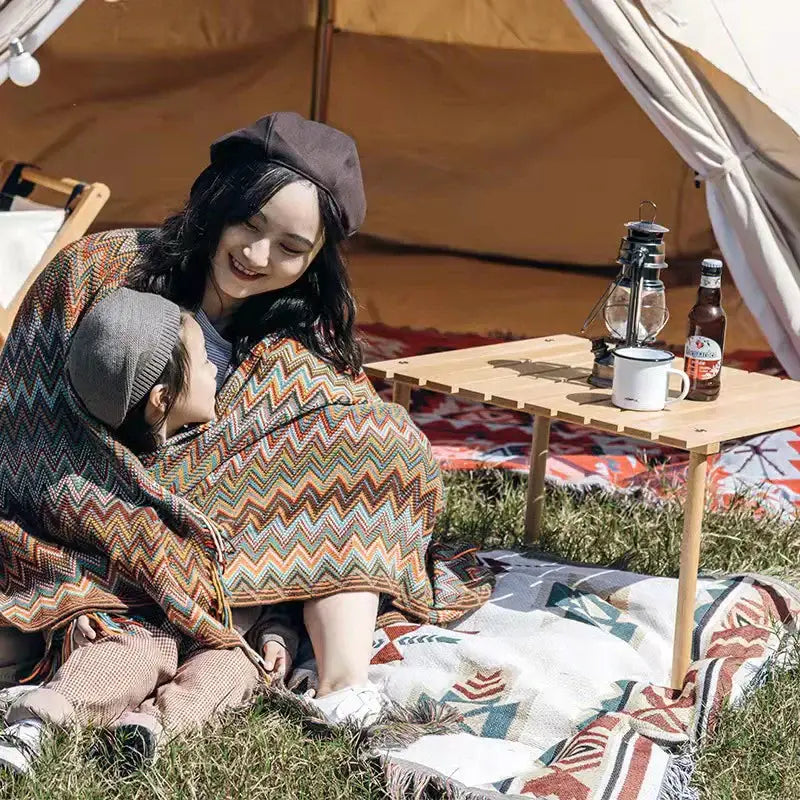 A cozy multicolored geometric aztec baja blanket in mustard and blue wrapped around woman and child with fringed edges