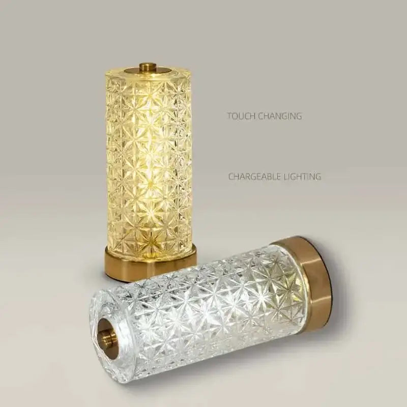 Two decorbites crystal table lamps with faceted glass bodies and brass caps, one glowing warmly amber, one clear