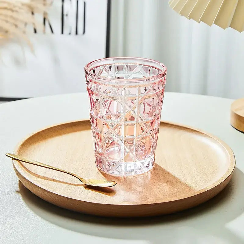 A pink faceted glass tumbler with a geometric pattern sits on a wooden tray as a heavy whiskey glass