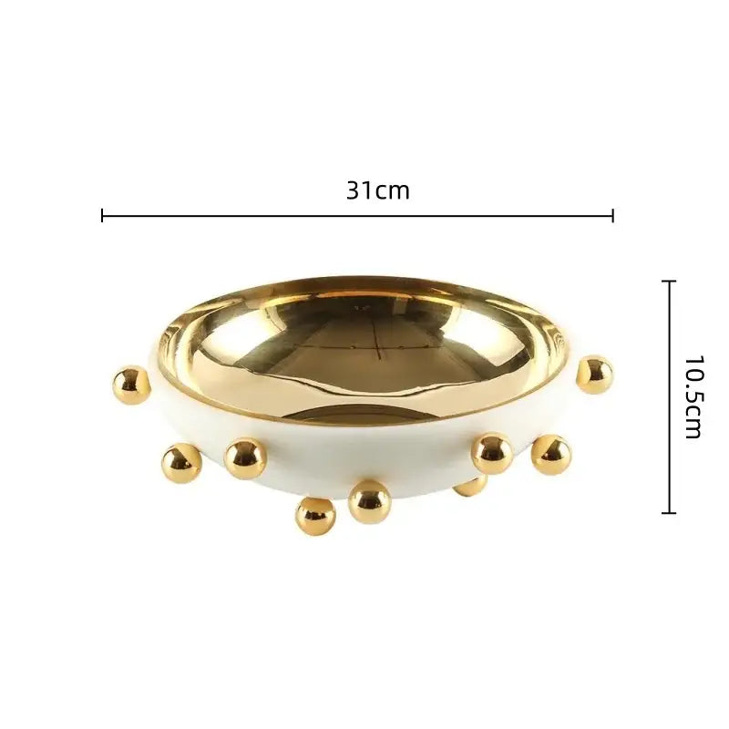 A sleek white ceramic fruit bowl round with polished gold interior and decorative gold ball accents on rim