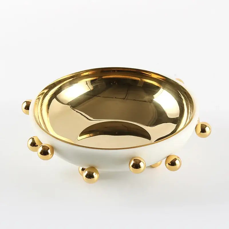 A sleek white ceramic fruit bowl round with polished gold interior and decorative gold ball accents on rim