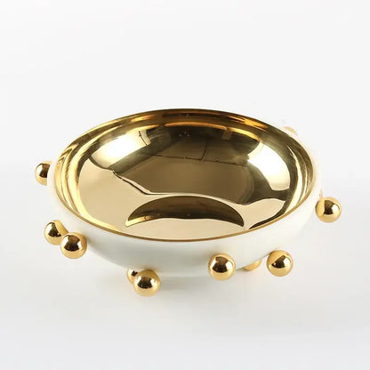 A sleek white ceramic fruit bowl round with polished gold interior and decorative gold ball accents on rim