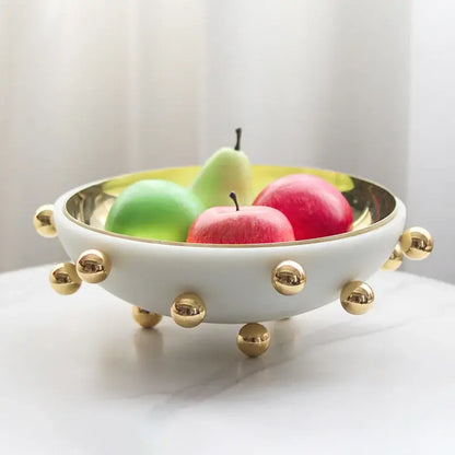 White ceramic fruit bowl round with gold-tone studs and reflective golden interior holding colorful apples and pears