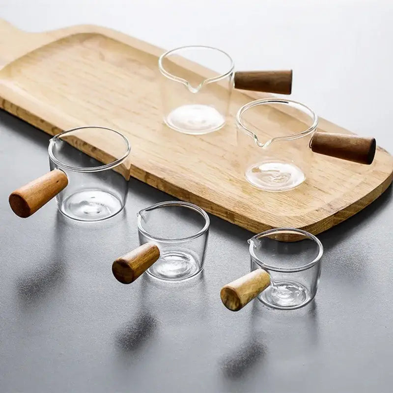 Set of four clear glass espresso measuring cups with wood handles arranged on a wooden tray for kitchen use