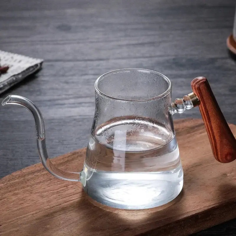 Clear glass goose beak kettle with polished wooden handle and brass accents filled with water for pour over coffee