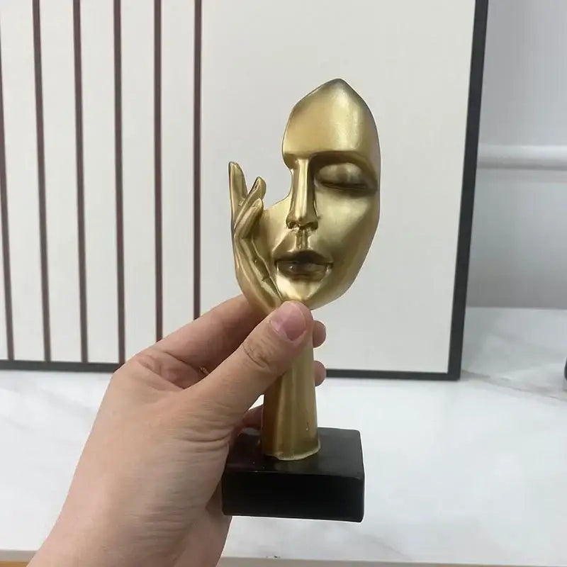 Golden metallic abstract mask statue with a hand touching its cheek mounted on a black base for home office decor