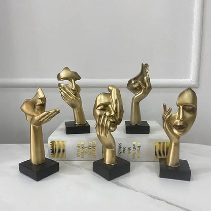 Golden abstract mask statue featuring metallic hand and face sculptures with sleek designs on black bases for decor