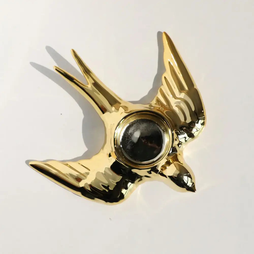 Golden swallow-shaped polished metallic fidget spinner shown as a decorative element on a golden bird ceramic candle holder tray