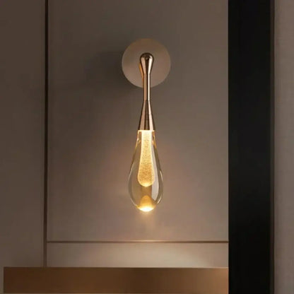 A sleek golden water droplets wall light with teardrop clear glass and illuminated filament bulb by decorbites
