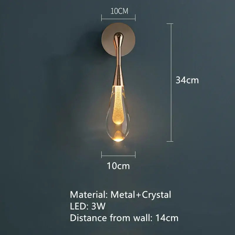 A sleek teardrop-shaped bedroom wall light with brass metal stem and clear crystal body with golden led light decorbites golden water droplets