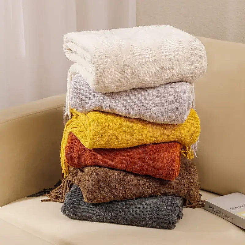 Stack of six decorbites knit blankets with cable knit patterns and fringed edges in cream, gray, yellow, orange, brown, and dark gray for sofa decor