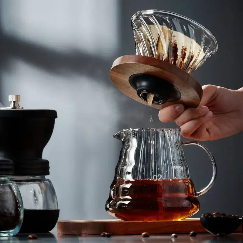 Clear glass coffee brewer with wooden base and black seal held above ribbed carafe in decorbites glass coffee set
