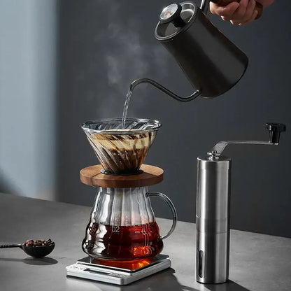 A sleek matte black gooseneck kettle with stainless steel spout pouring water over a decorbites glass coffee brewer