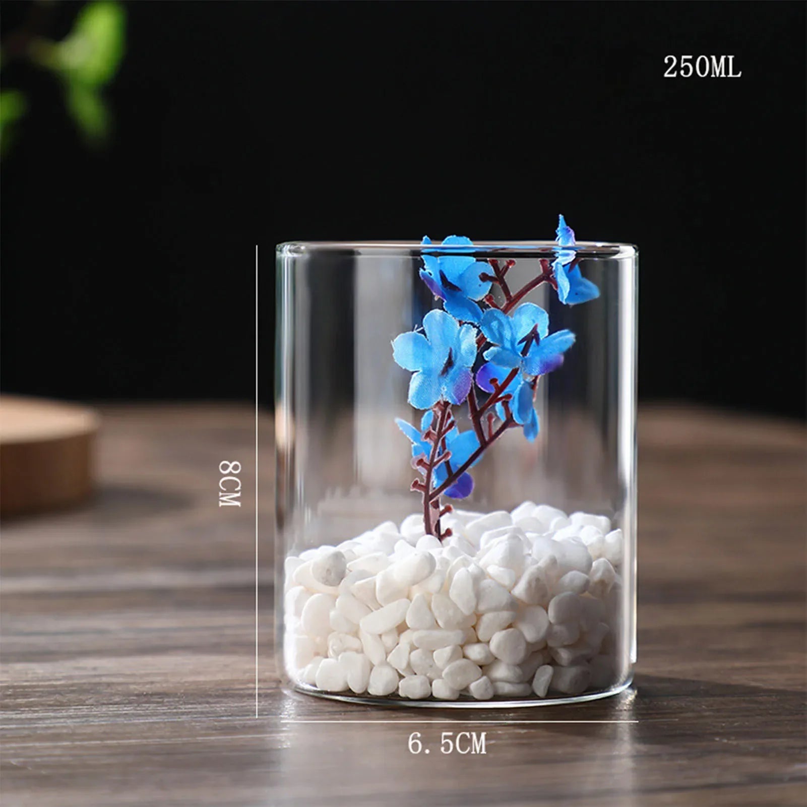 Clear glass cylindrical hydroponics planter vase filled with white pebbles and blue artificial flowers for desktop decor