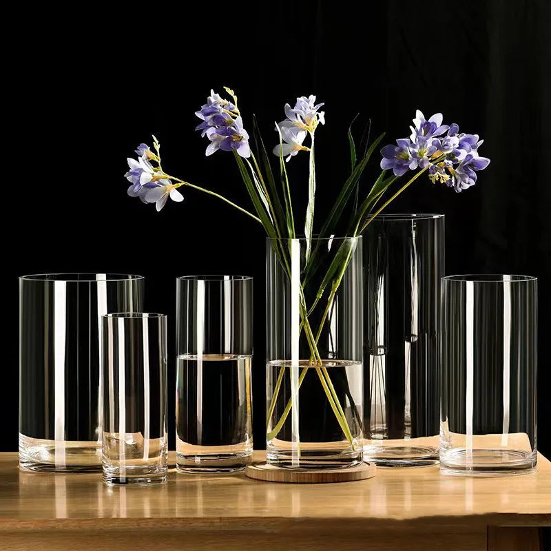 Clear cylindrical glass vases with subtle vertical ridges and one holding purple flowers on wooden coaster for desktop decor