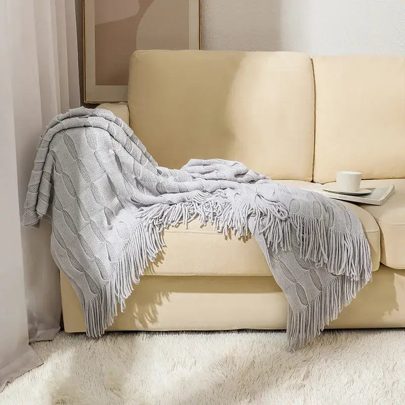A soft light gray decorbites knitted geometric throw blanket with cable knit and fringe draped over a beige sofa