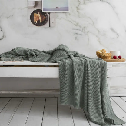 A soft sage green knitted bamboo cooling throw blanket with textured weave draped over a white wooden bench