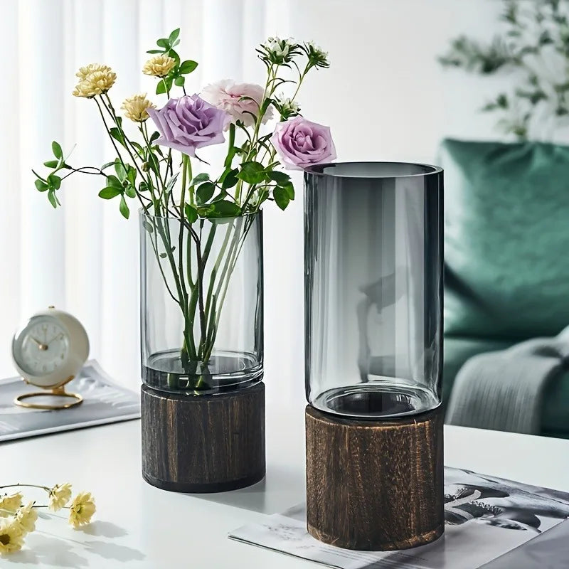 Two cylindrical glass vases with dark wood bases, one containing purple and yellow flowers, decorative for living room table