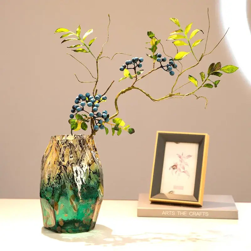 A marbled green and black glass vase with swirling gold gradients black glass holds branches with blue berries for decorbites gradients black