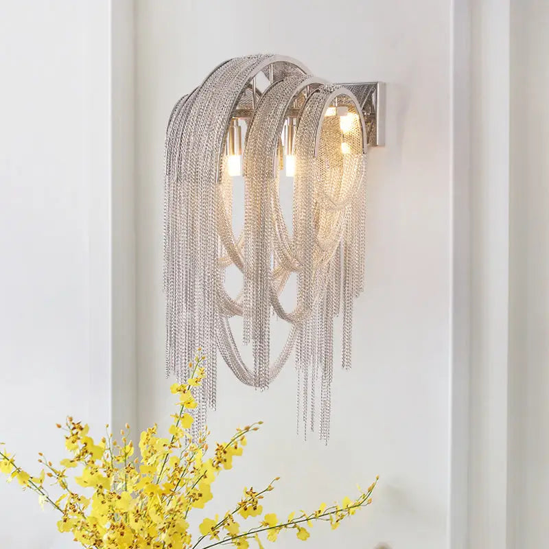 Silver stacked tassel wall lamp with cascading metallic strands and three illuminated bulbs for bedroom sconce lighting