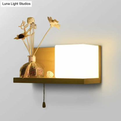 Gold-toned wall light with milk glass cube rectangular panel and wood shelf featuring pull-chain switch for bedroom use