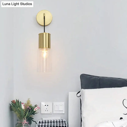 Minimal brass and glass wall sconce light with gold-toned cylindrical arm and clear glass shade for bedroom use
