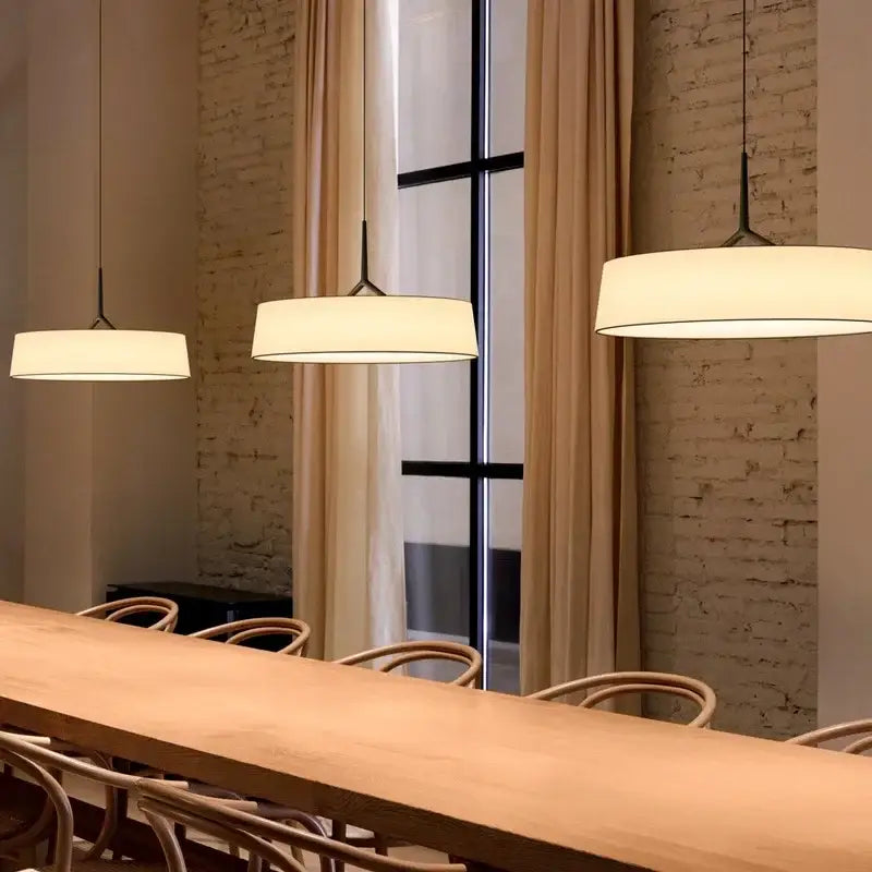 Three minimalist dama pendant lamps with white fabric shades and black metal fixtures hang above a wooden table