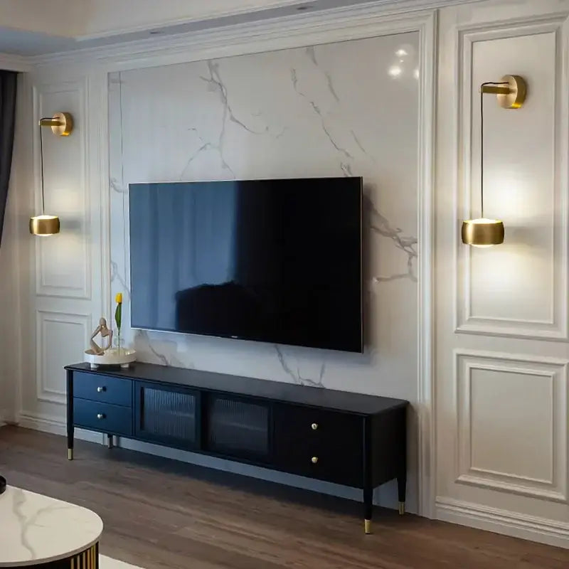 Black tv stand with gold-tone legs silver knobs and glass-fronted cabinets paired with minimalist brushed gold wall lamp
