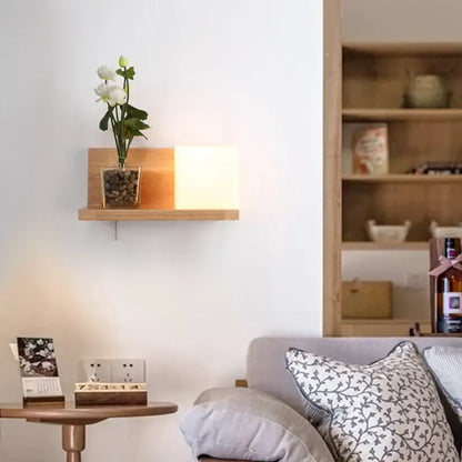 Minimalist frosted glass wooden wall lamp with pull chain beside a wooden wall shelf holding white flowers and pebbles