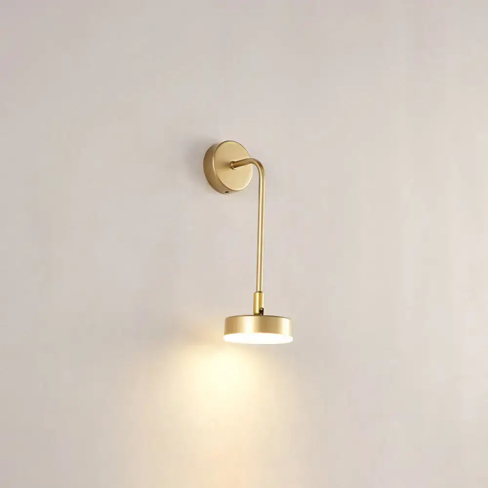 A minimalist gold metal led sconce wall mounted lamp with curved arm and circular base emitting warm light for bedroom