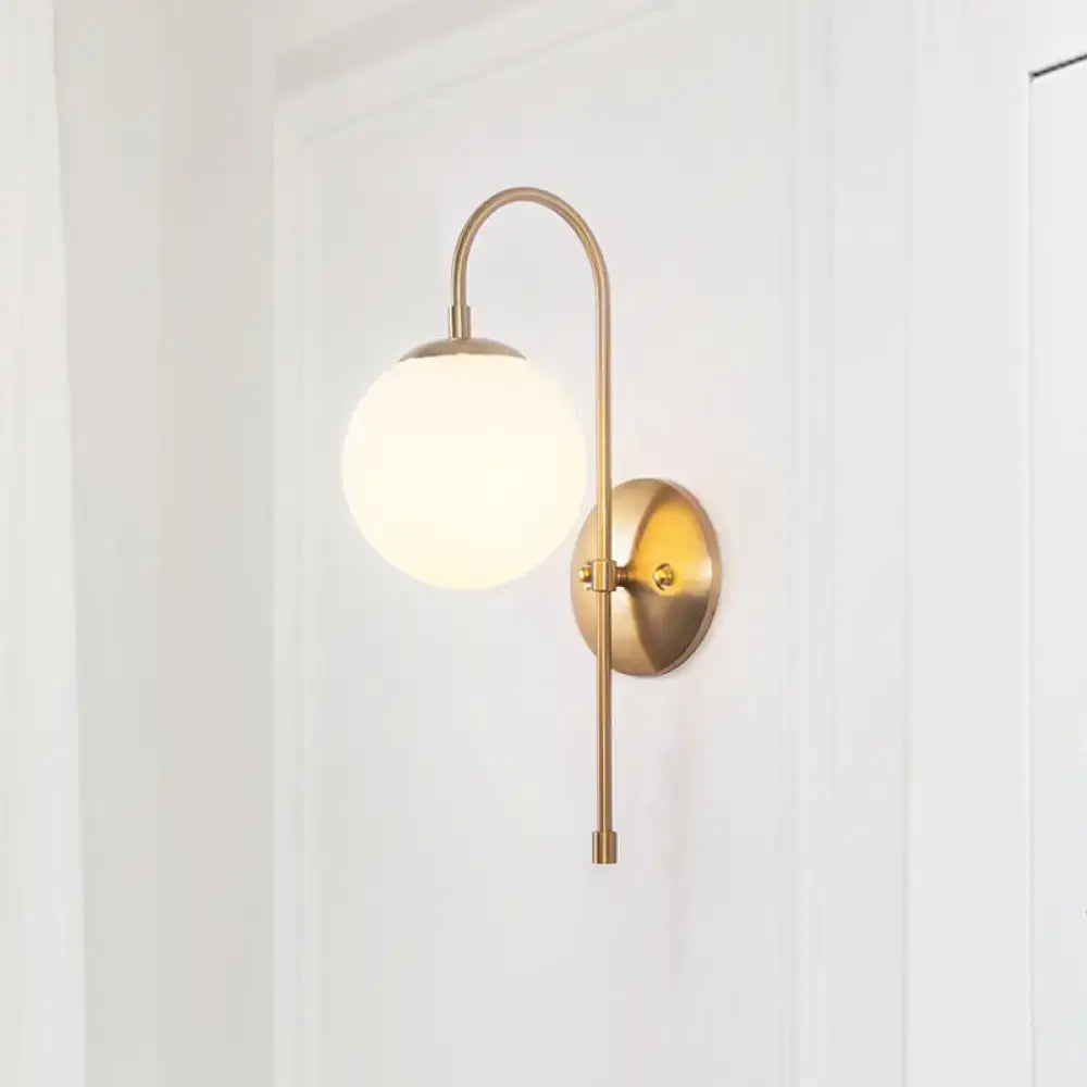 A sleek minimalist gold mini globe wall sconce with a curved brass arm and frosted glass globe shade for bedroom lighting